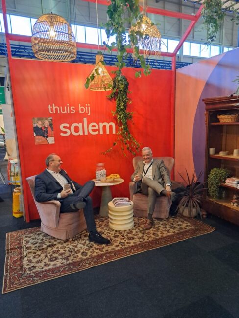 Twee mannen zitten in comfortabele stoelen bij een stand met de tekst "thuis bij salem", omringd door planten en decor.