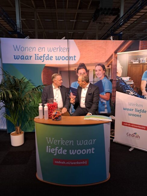 Twee mannen zitten aan een tafel met promotiemateriaal van Cedrah, met de tekst "Wonen en werken waar liefde woont".