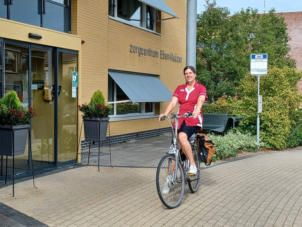 Vrouw in rood uniform fietst voor een zorgcentrum met gele bakstenen en groene planten bij de ingang op een zonnige dag.