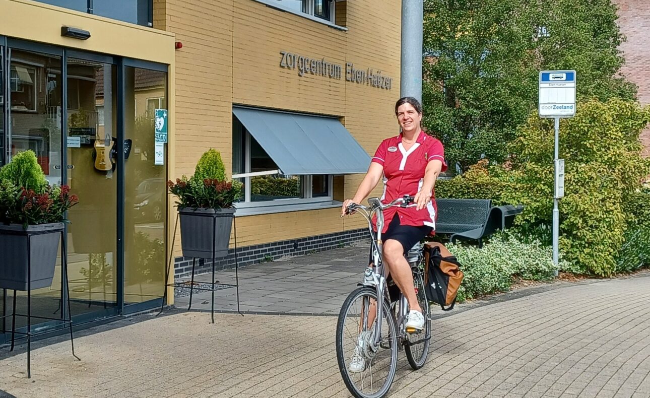 Vrouw in rood uniform fietst voor een zorgcentrum met gele bakstenen en groene planten bij de ingang op een zonnige dag.