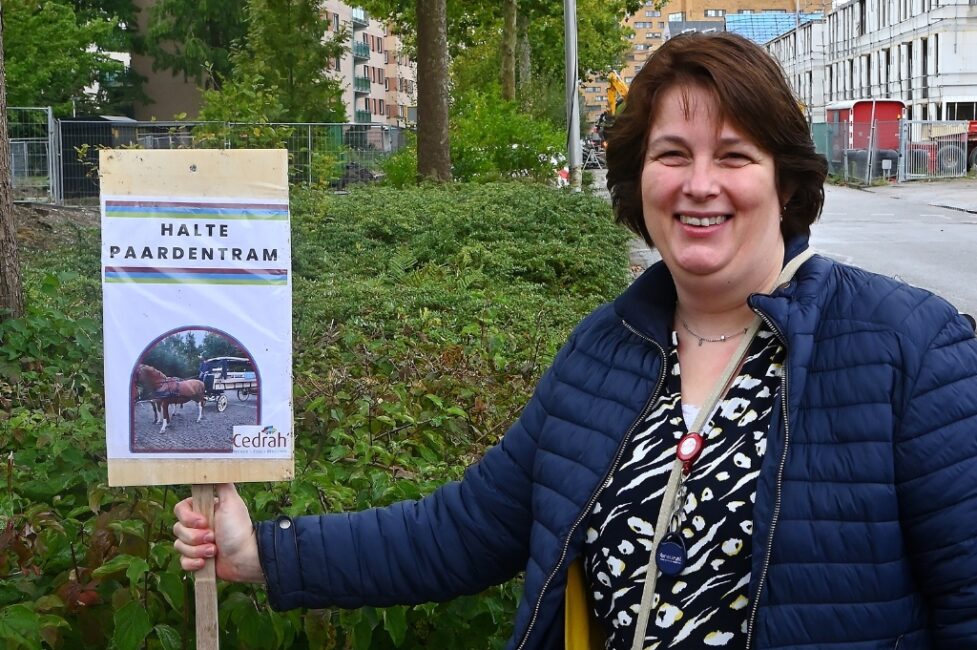 Vrouw met blauwe jas lacht naast een bord met de tekst "Halte Paardentram" en een afbeelding van een paardentram.