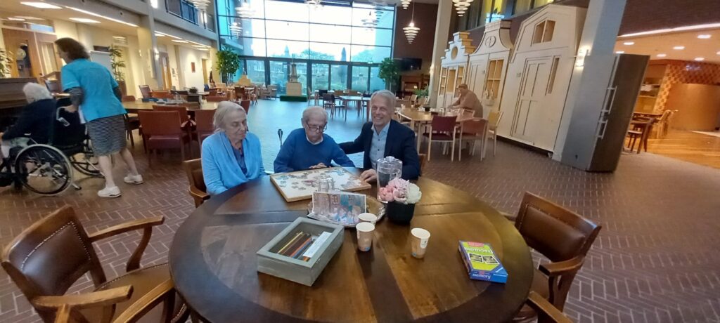 Drie oudere mensen zitten aan een ronde tafel in een gemeenschappelijke ruimte, omringd door boeken en koffiekopjes.