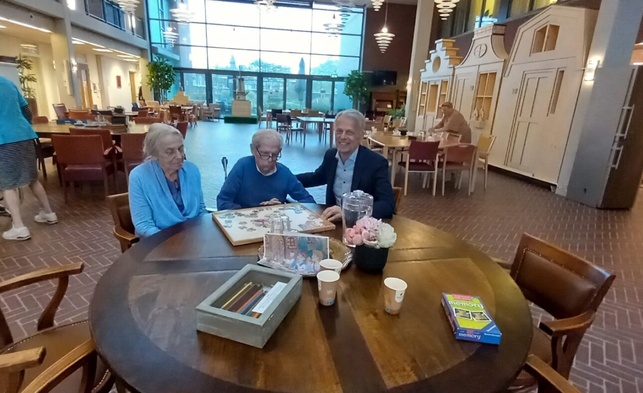 Drie oudere mensen zitten aan een ronde tafel in een gemeenschappelijke ruimte, omringd door boeken en koffiekopjes.
