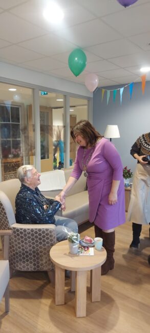 Een vrouw in een paarse jurk schudt de hand van een oudere man die in een stoel zit in een gezellige kamer met ballonnen.