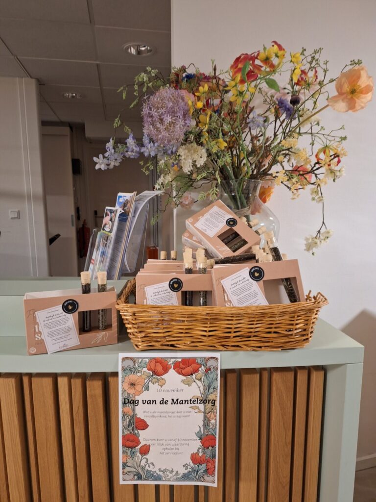 Een mand met cadeaus en een bloemenvaas op een tafel. Er is een poster voor de Dag van de Mantelzorg zichtbaar.