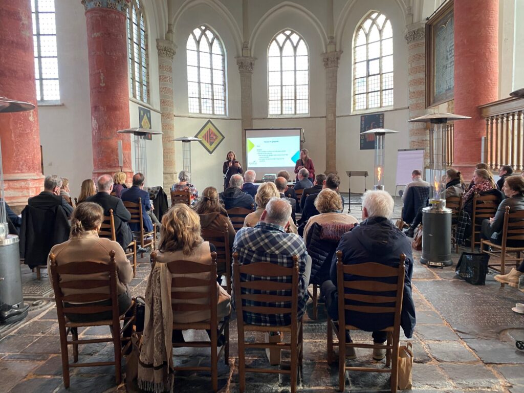 Groep mensen zit op stoelen in een historische zaal met hoge ramen, tijdens een presentatie op een themadag voor leidinggevenden.