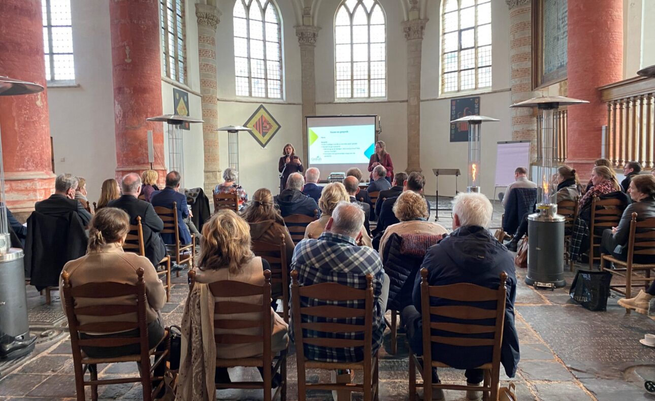 Groep mensen zit op stoelen in een historische zaal met hoge ramen, tijdens een presentatie op een themadag voor leidinggevenden.