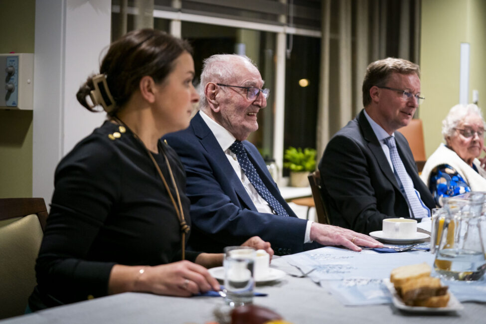 Vier mensen zitten aan een tafel met drinken en broodjes. Ze zijn in gesprek in een verlichte kamer met gordijnen.