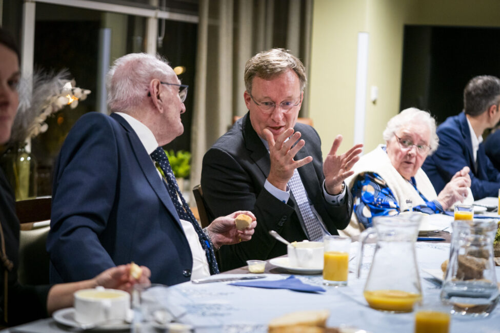 Een groep mensen zit aan een gedekte tafel tijdens een bijeenkomst. Ze zijn in gesprek en er staan glazen met sinaasappelsap.