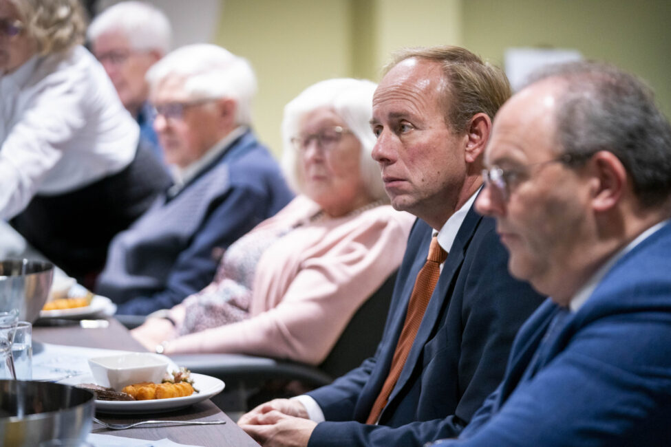 Groep mensen zit aan een tafel tijdens een bijeenkomst. Een man in een pak kijkt aandachtig. Er staan schalen met eten op tafel.