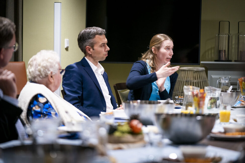 Een groep mensen zit aan een tafel met eten en drinken, in gesprek. Een vrouw gebaart terwijl ze iets uitlegt.