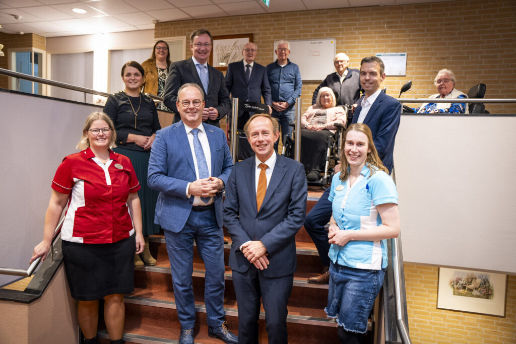 Groepsfoto van mensen op een trap in een gebouw, sommigen in uniform. Ze lachen en poseren voor de camera.
