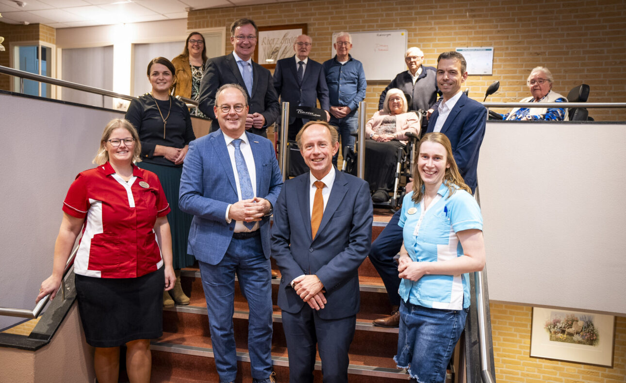 Groepsfoto van mensen op een trap in een gebouw, sommigen in uniform. Ze lachen en poseren voor de camera.