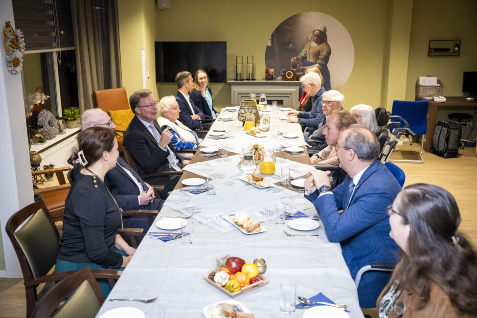 Groep mensen zit aan een lange tafel in een vergaderruimte. Er zijn drankjes en fruit op tafel. Iedereen luistert aandachtig.