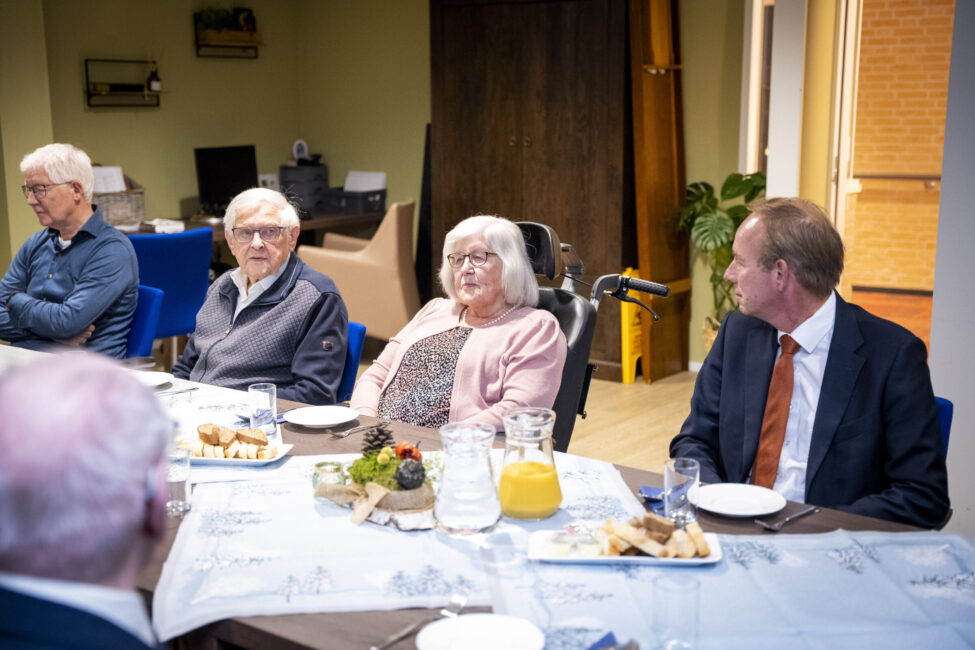 Groep mensen zit aan een tafel met eten en drinken in een gemeenschappelijke ruimte. Ze zijn in gesprek met elkaar.