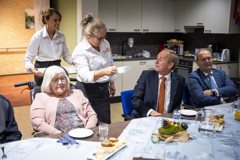 Groep mensen zit aan een tafel in een eetzaal. Een serveerster biedt een kopje koffie aan een man in een pak aan.