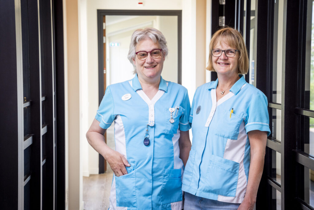 Twee verpleegkundigen in blauwe uniformen staan glimlachend in een gang van een zorginstelling, met deuren aan beide zijden.