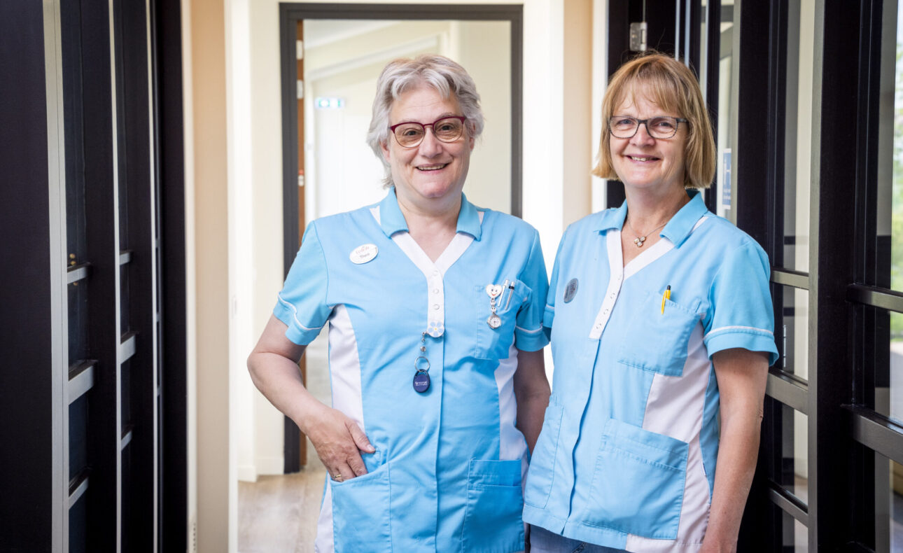Twee verpleegkundigen in blauwe uniformen staan glimlachend in een gang van een zorginstelling, met deuren aan beide zijden.