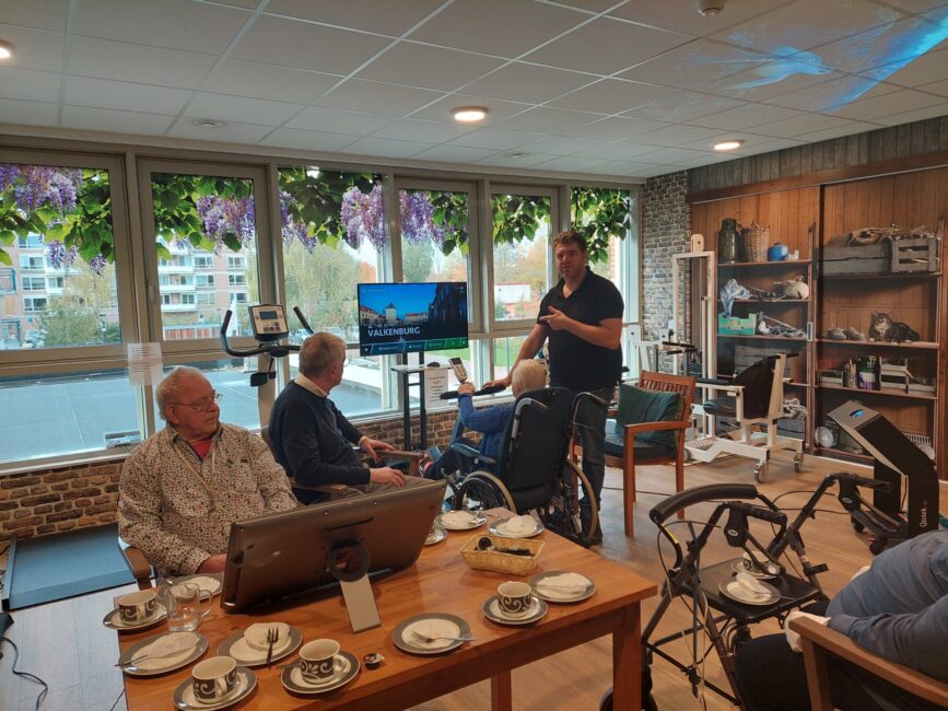 Mensen in een kamer met een groot scherm en fietsen. Er zijn tafels met kopjes en een man geeft uitleg aan de groep.