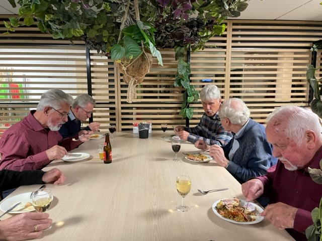 Zes oudere mannen zitten aan een tafel en eten samen een maaltijd in een gezellige ruimte met houten lambrisering en planten.