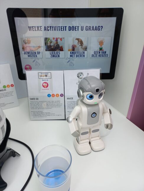 Een kleine witte robot staat op een tafel voor een scherm met de tekst "Welke activiteit doet u graag?" en enkele opties.