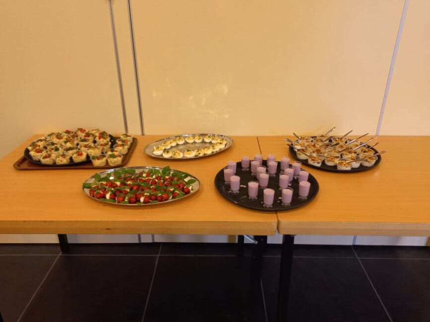 Een tafel met diverse hapjes, waaronder gevulde eieren, spiesjes met tomaat en mozzarella, en paarse dessertbekers.