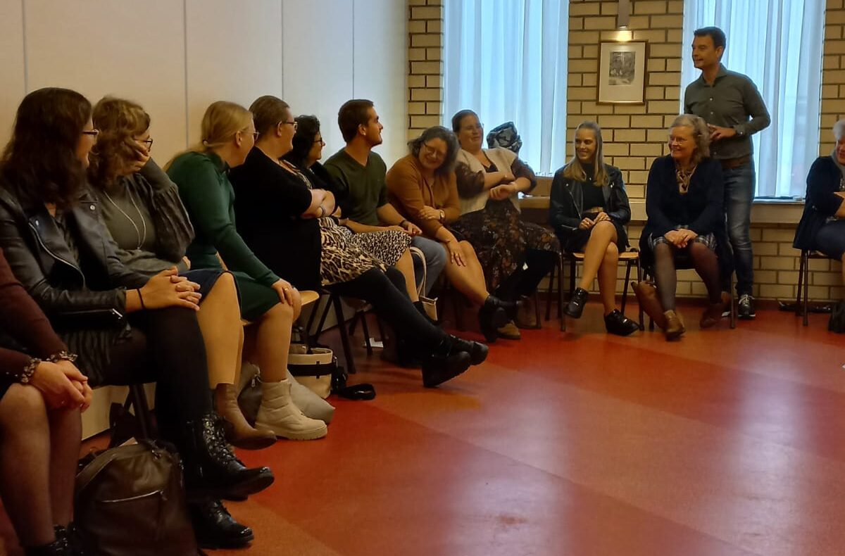 Groep mensen zit in een kring in een zaal met rode vloer en bakstenen muur. Een persoon staat en spreekt tot de groep.
