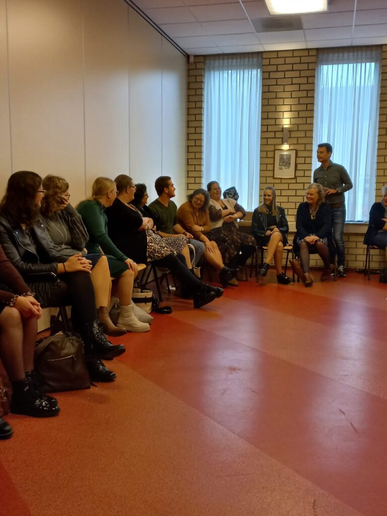 Groep mensen zit in een kring in een zaal met rode vloer en bakstenen muur. Een persoon staat en spreekt tot de groep.