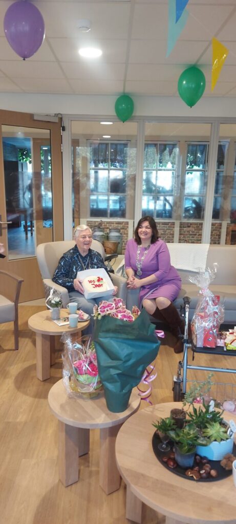 Twee vrouwen zitten in een feestelijk versierde kamer met ballonnen. Een van hen houdt een taart vast. Er zijn cadeaus op tafel.