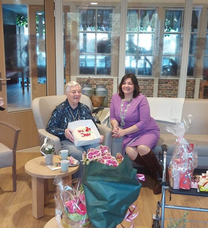 Twee vrouwen zitten in een feestelijk versierde kamer met ballonnen. Een van hen houdt een taart vast. Er zijn cadeaus op tafel.