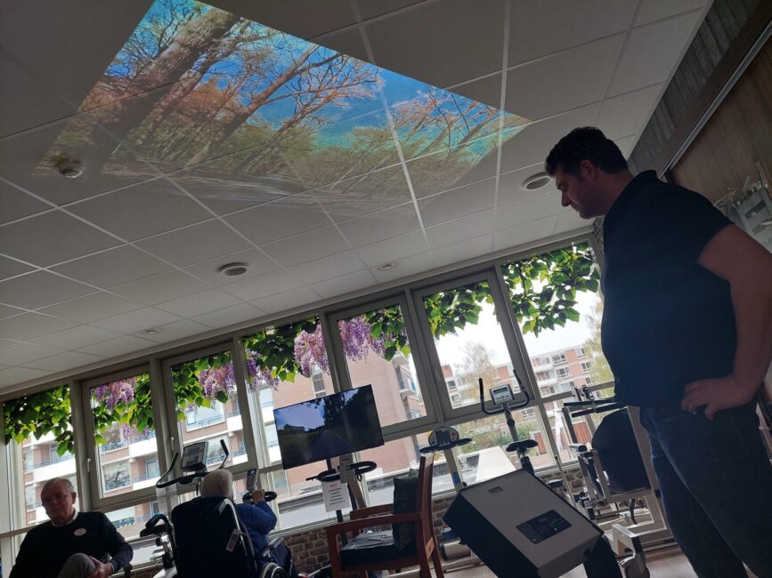 Een man kijkt naar een plafondprojectie van een boslandschap in een kamer met grote ramen en fitnessapparatuur.