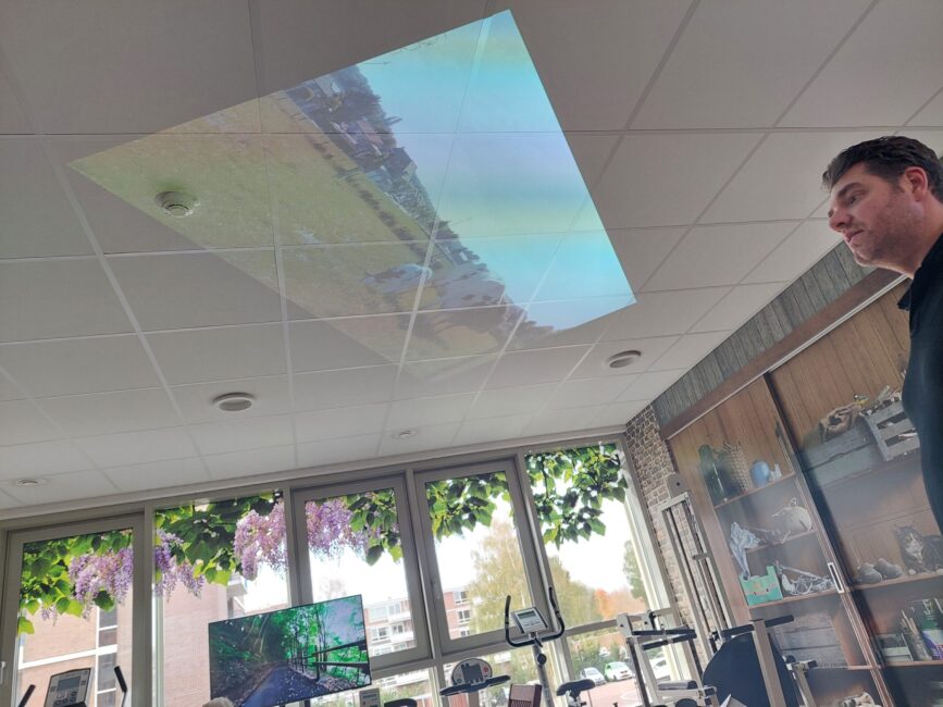 Man kijkt naar een plafondprojectie van een landschap in een moderne ruimte met grote ramen en fitnessapparatuur.