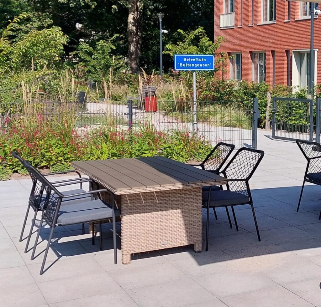 Tuinmeubilair op een zonnig terras met een tafel en stoelen, omgeven door groene planten en een bakstenen gebouw op de achtergrond.