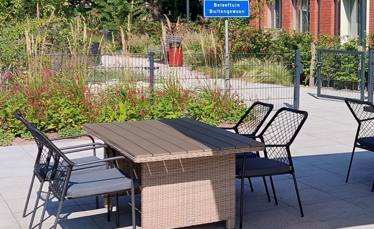 Tuinmeubilair op een zonnig terras met een tafel en stoelen, omgeven door groene planten en een bakstenen gebouw op de achtergrond.