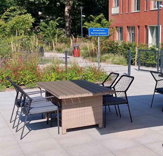 Tuinmeubilair op een zonnig terras met een tafel en stoelen, omgeven door groene planten en een bakstenen gebouw op de achtergrond.