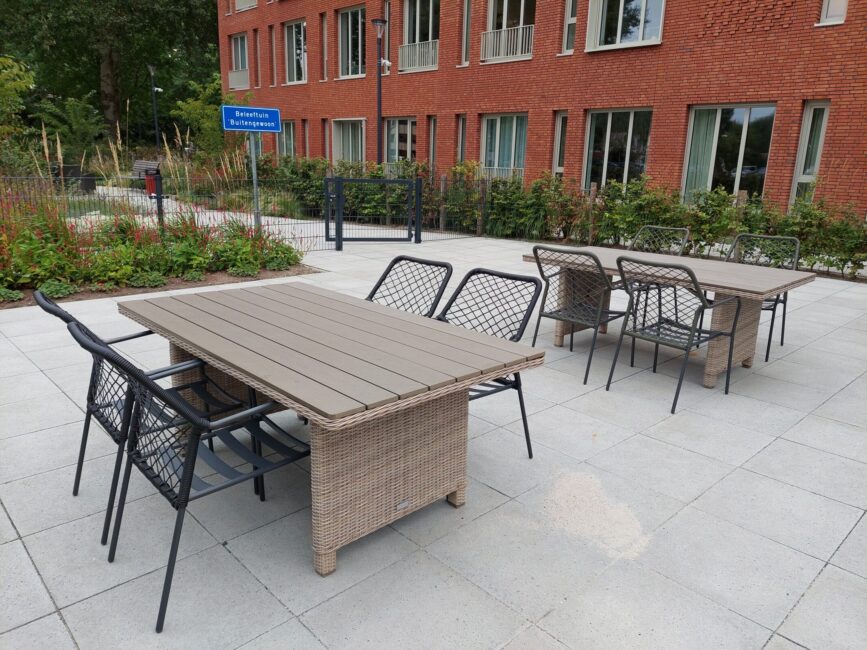 Tuinmeubilair op een betegeld terras met twee tafels en acht stoelen, omgeven door groen en een bakstenen gebouw op de achtergrond.