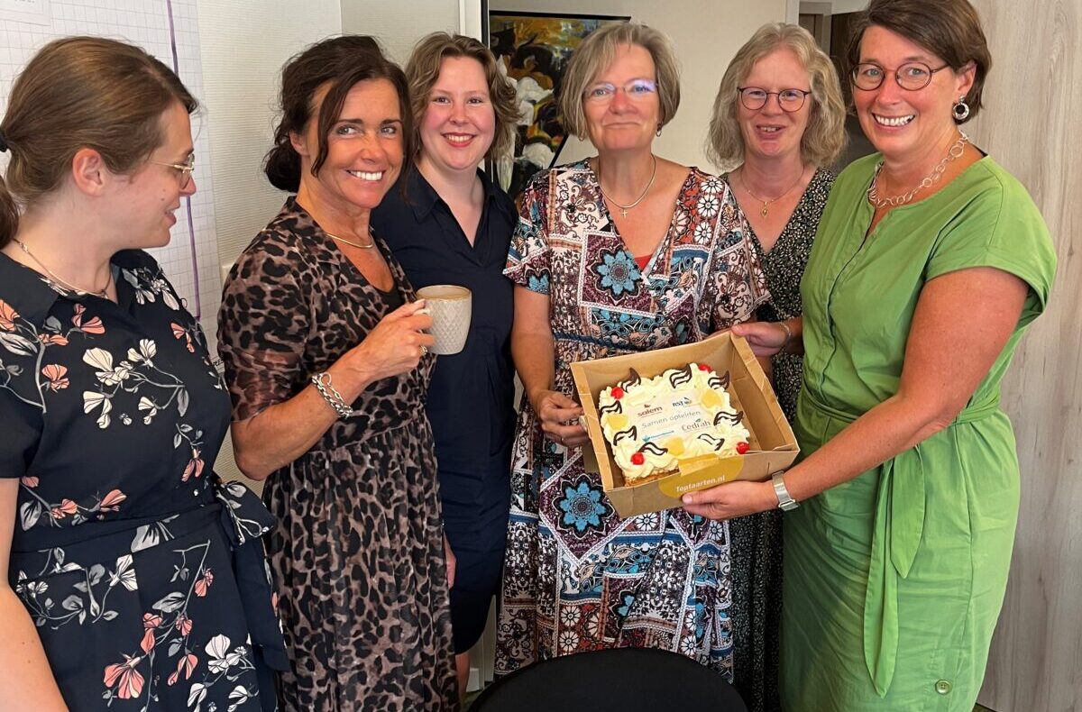 Een groep van zes lachende vrouwen staat in een kamer. Eén van hen houdt een doos met gebak vast. Ze lijken een feestje te vieren.
