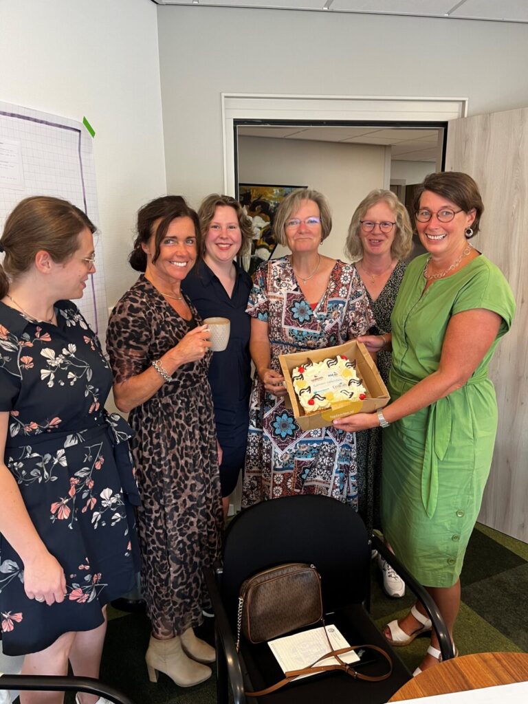 Een groep van zes lachende vrouwen staat in een kamer. Eén van hen houdt een doos met gebak vast. Ze lijken een feestje te vieren.