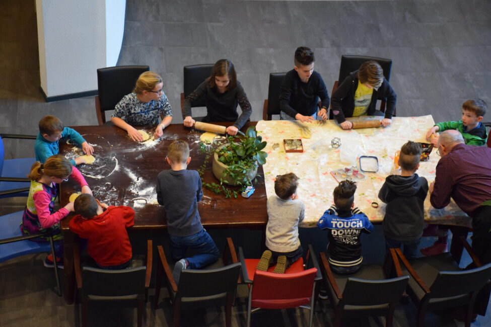 Groep kinderen en volwassenen bakt koekjes aan een grote tafel, omringd door stoelen, met bloem en deeg op het tafelblad.