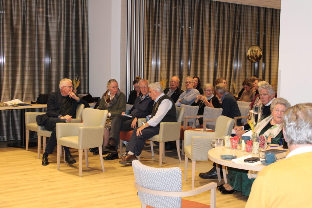 Groep oudere mensen zit in een zaal met gordijnen, luisterend naar een presentatie. Tafels met kopjes en glazen zijn zichtbaar.