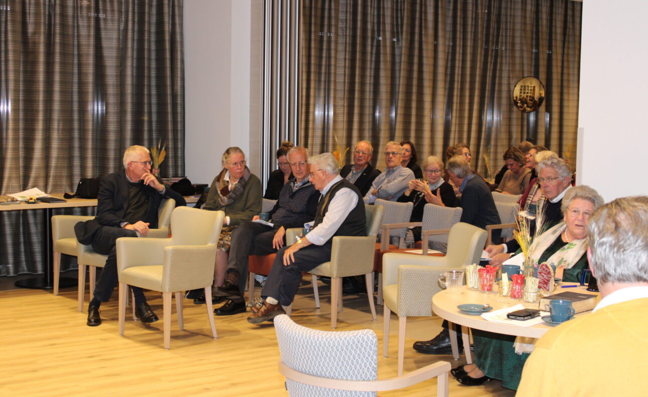 Groep oudere mensen zit in een zaal met gordijnen, luisterend naar een presentatie. Tafels met kopjes en glazen zijn zichtbaar.