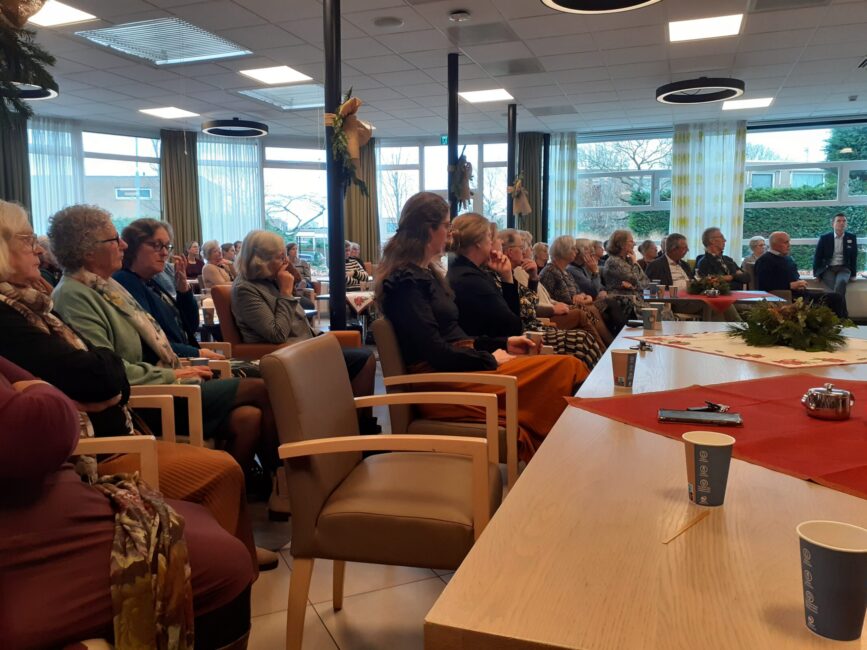 Groep mensen zit in een gemeenschappelijke ruimte, luisterend naar een presentatie. Tafels zijn gedekt met rode kleden en kopjes.