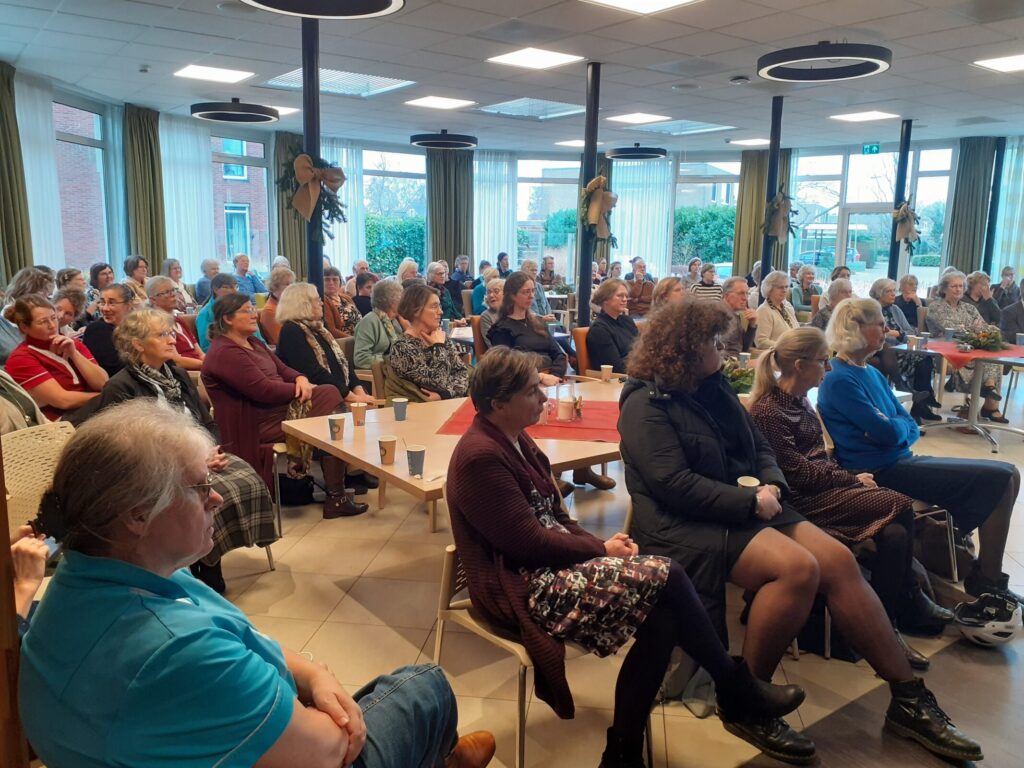 Een groep mensen zit in een zaal met tafels en stoelen, aandachtig luisterend naar een presentatie of spreker.