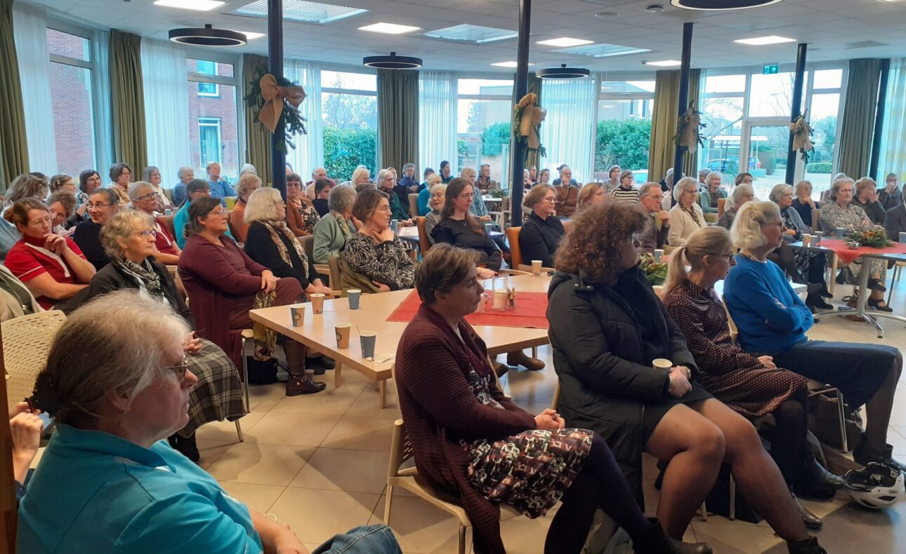 Een groep mensen zit in een zaal met tafels en stoelen, aandachtig luisterend naar een presentatie of spreker.