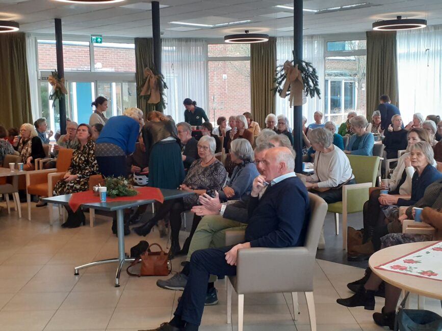 Groep mensen zit in een gemeenschappelijke ruimte met tafels en stoelen, bezig met een sociale activiteit of bijeenkomst.