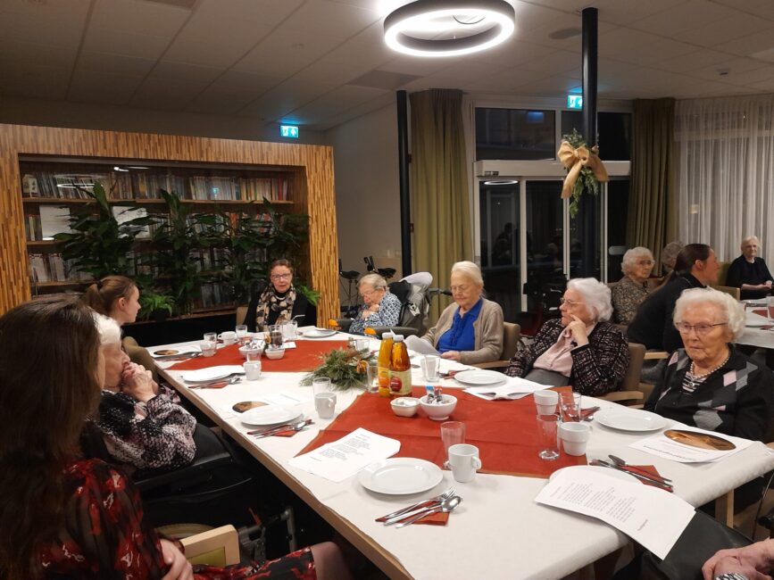 Een groep ouderen zit aan een feestelijk gedekte tafel in een gezellige ruimte, met kerstversiering en warme verlichting.