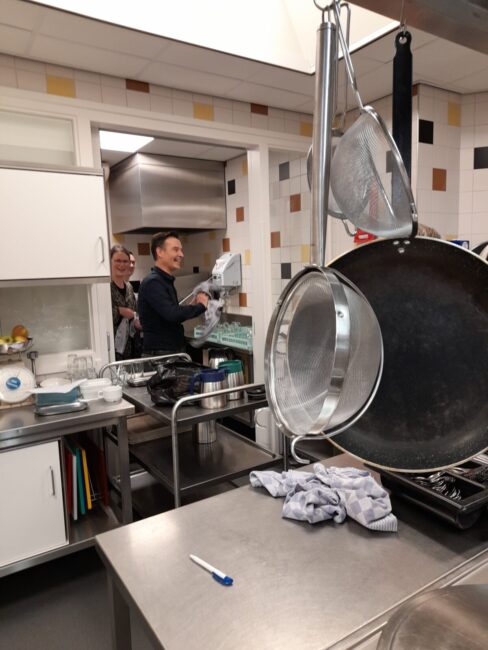 Twee mensen in een keuken, bezig met afwassen. Op de voorgrond hangen pannen en liggen doeken op het aanrecht.