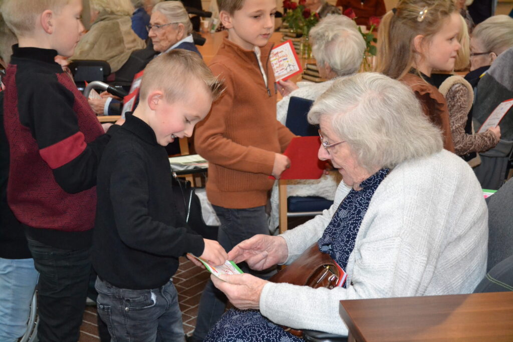 Kinderen geven kaarten aan een oudere vrouw in een drukke ruimte. De vrouw glimlacht terwijl ze de kaarten bekijkt.
