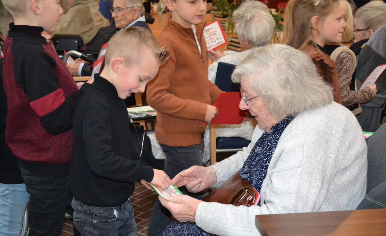 Kinderen geven kaarten aan een oudere vrouw in een drukke ruimte. De vrouw glimlacht terwijl ze de kaarten bekijkt.
