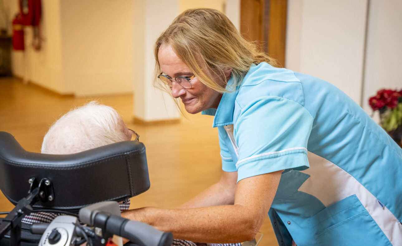 Een zorgmedewerker in een blauw uniform buigt zich glimlachend naar een oudere persoon in een rolstoel in een lichte kamer.